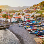 Porto colorato di Madeira con barche tradizionali ormeggiate sulla spiaggia di ciottoli, case bianche e verdeggianti colline terrazzate al tramonto, perfetto per viaggi e turismo