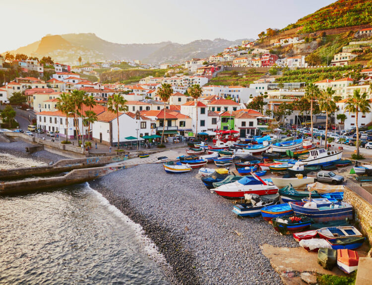 Porto colorato di Madeira con barche tradizionali ormeggiate sulla spiaggia di ciottoli, case bianche e verdeggianti colline terrazzate al tramonto, perfetto per viaggi e turismo