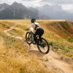 Biker che percorre un sentiero di montagna al Bike Park di Plan de Corones durante l'autunno, circondato da prati e alberi con fogliame dai colori caldi, con montagne sullo sfondo.